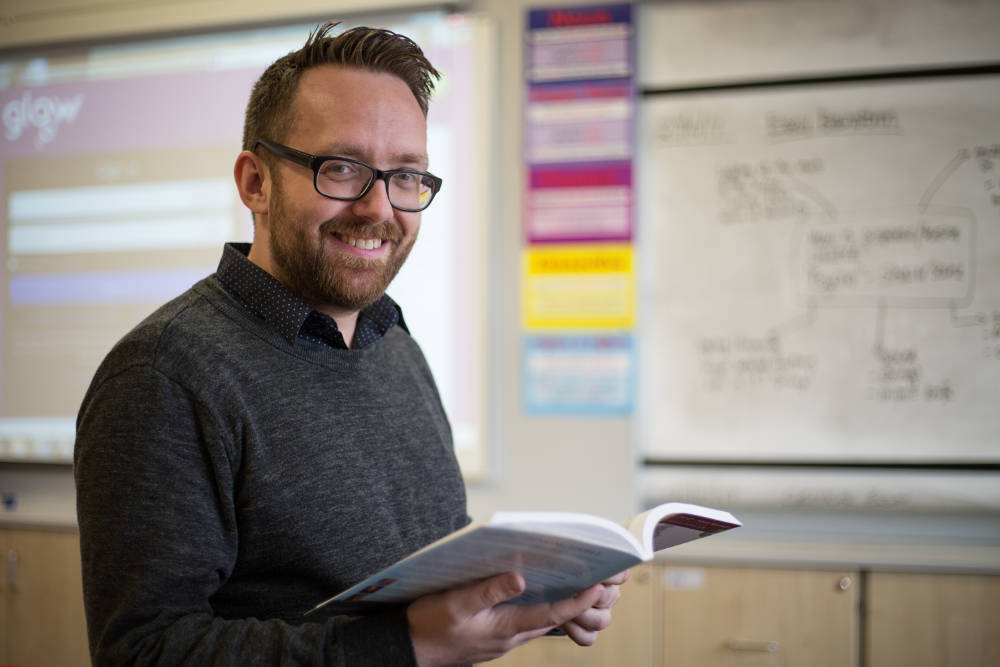 PGDE Teacher | Male holding open book in front of whiteboard